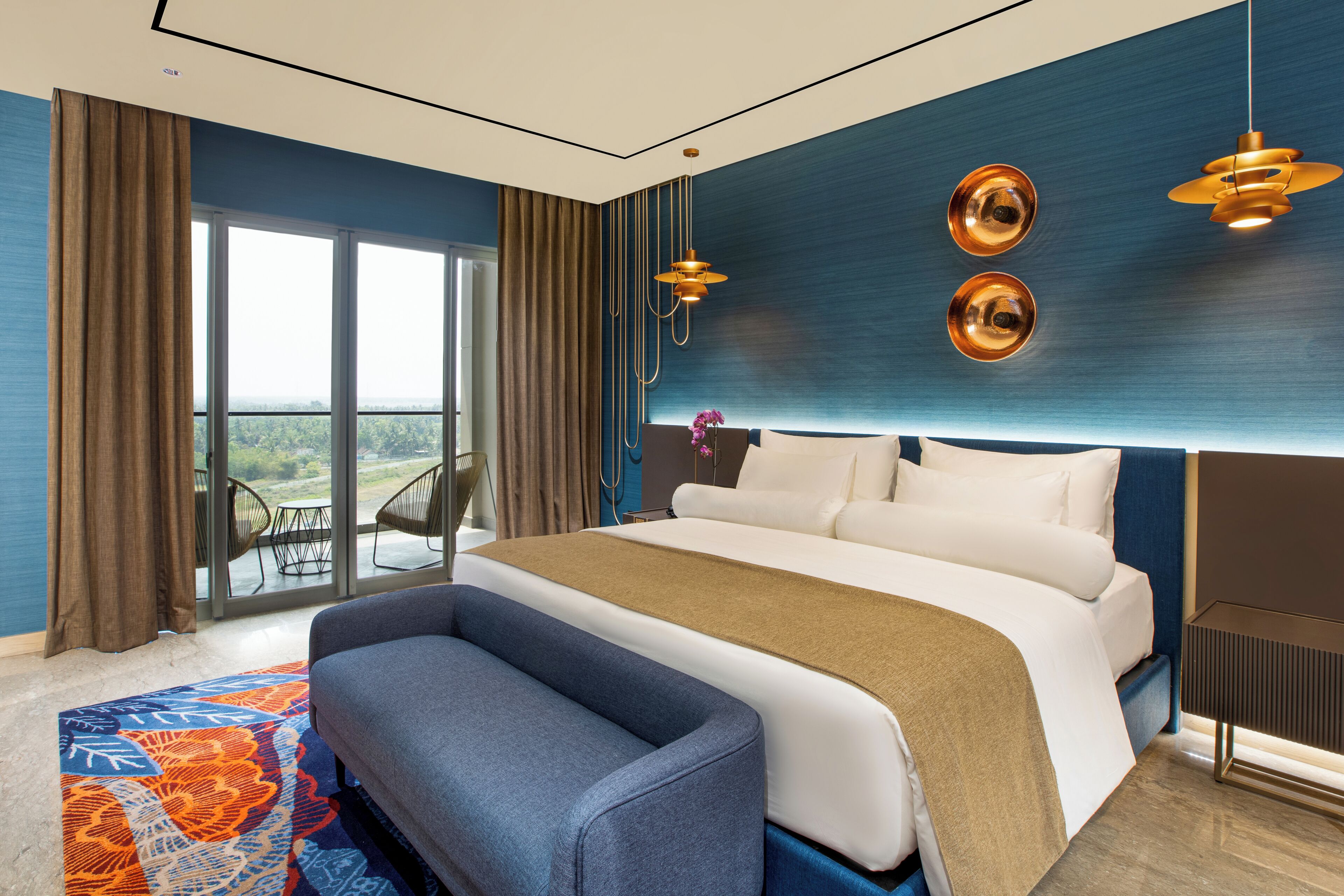 A modern hotel room with a large bed, a bench, a view through the window, and a blue accent wall.