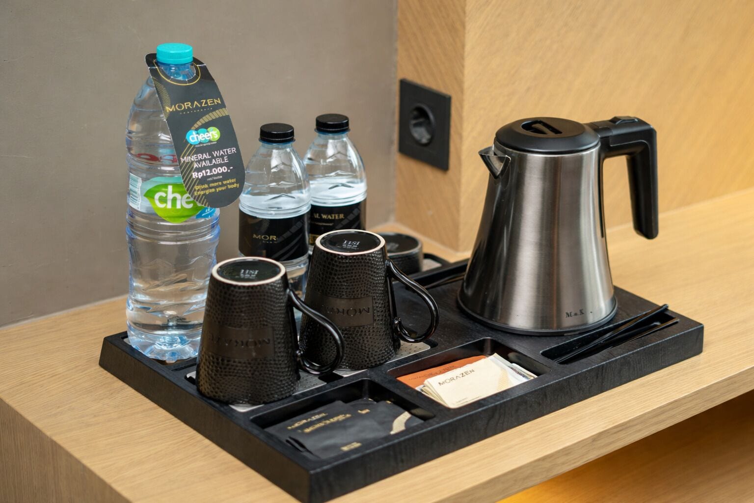 A hotel minibar setup with a water bottle, multiple glasses, a coffee pot, and a tray with a card.