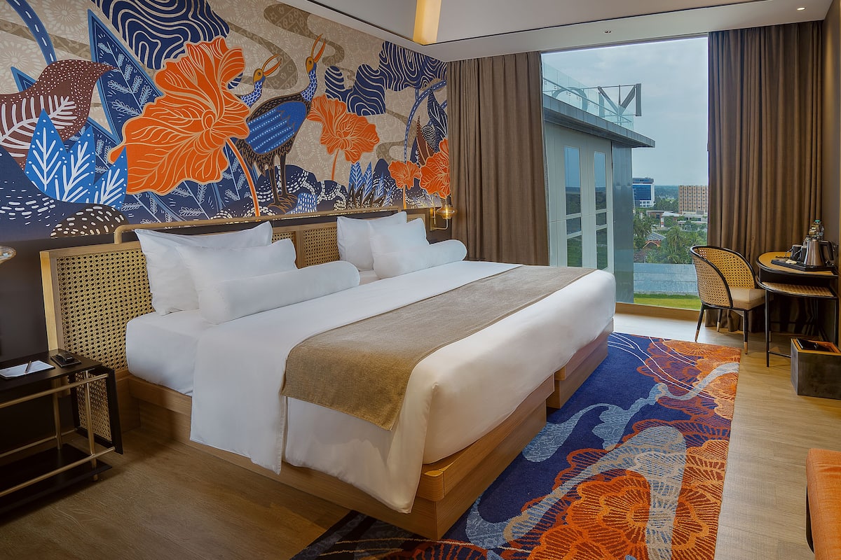 A hotel room with a large bed, a desk with a chair, a view of the city, and a decorative wall mural.