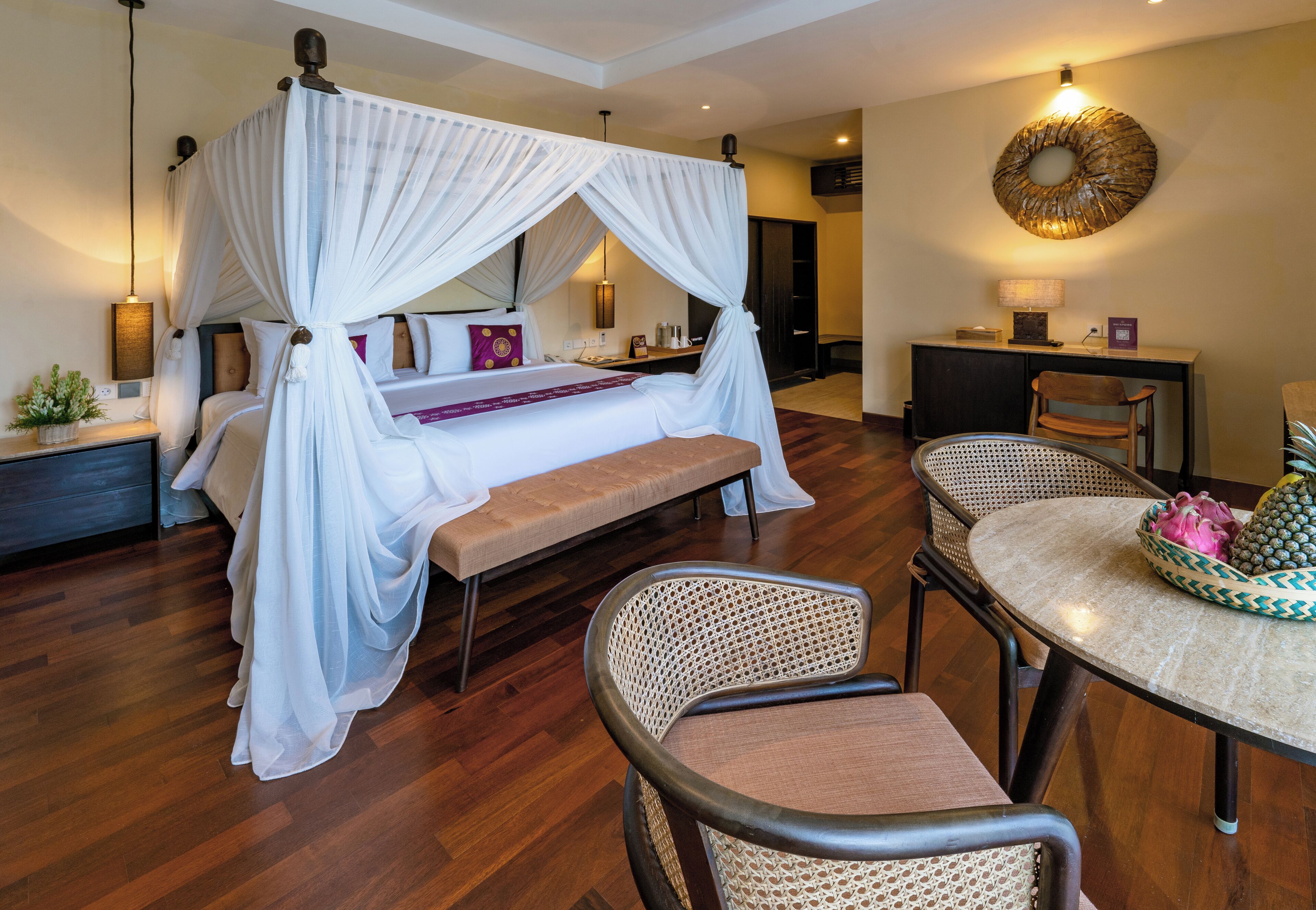 A bedroom with a four-poster bed, a bedside table, a chair, a round table, and a decorative wall piece.