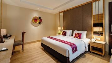 A hotel room with a large bed, a desk with a chair, a bedside table with a lamp, and a mirror on the wall.