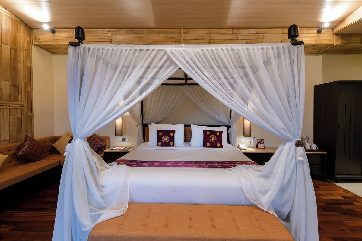 A bedroom with a canopy bed, a wooden headboard, a bedside table with a lamp, and a nightstand with a phone and a decorative item.