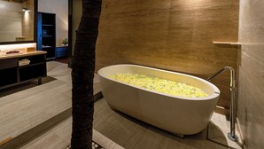 Un baño moderno con una bañera independiente llena de flores amarillas, una maceta con palmera y una pared de madera.