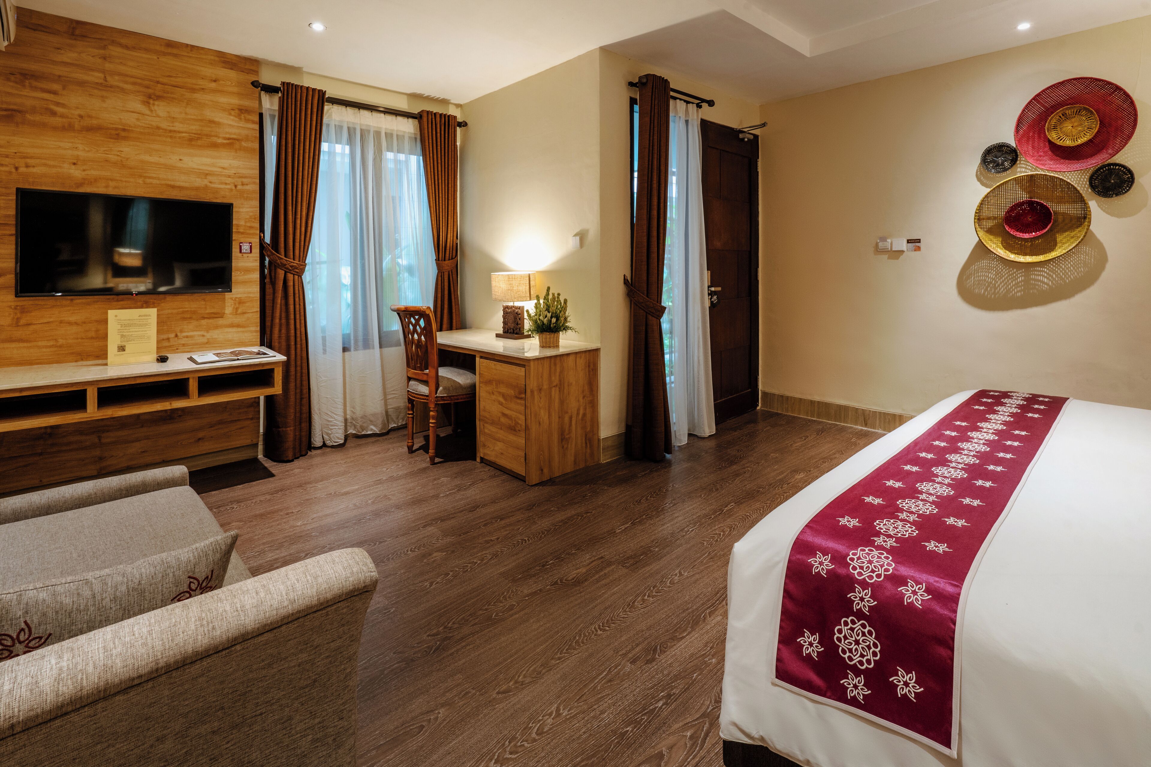Ein Hotelzimmer mit Holzboden, einem Bett mit rotem und weißen Überwurf, einem Schreibtisch mit Lampe, einem an der Wand befestigten Fernseher und einem großen Fenster mit Vorhängen.