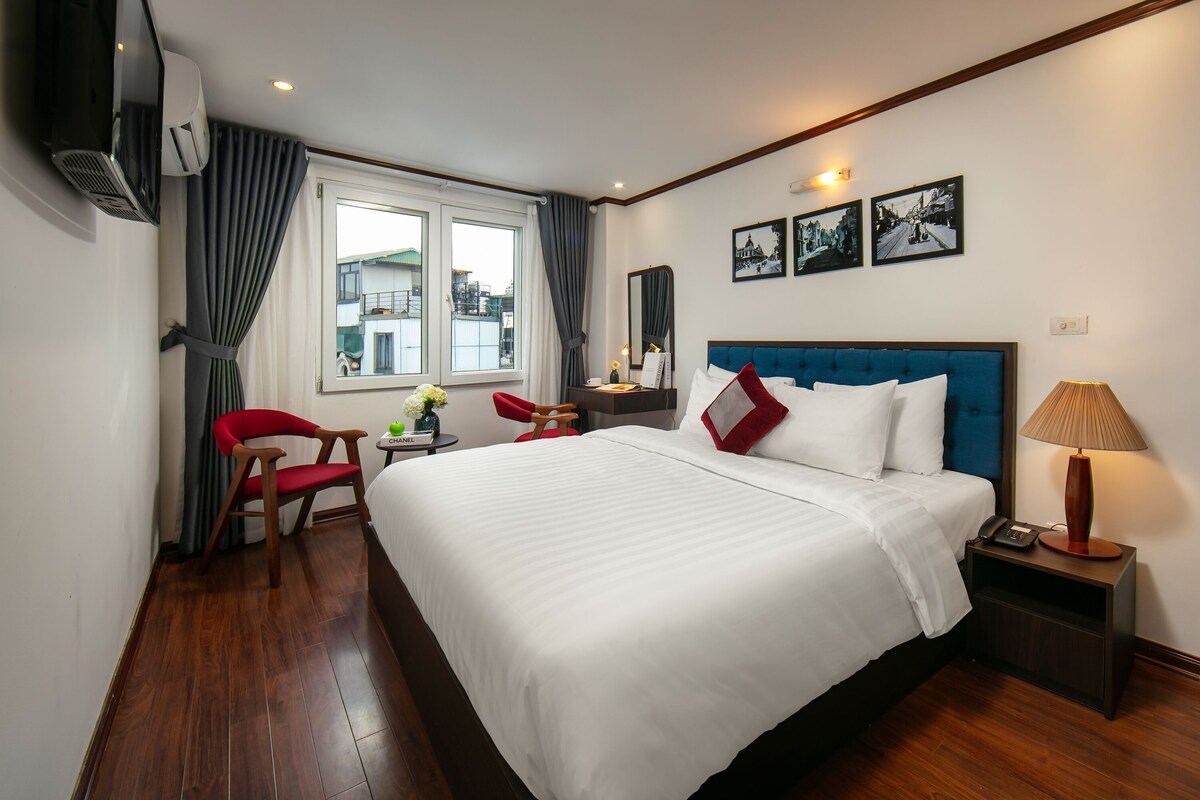 A hotel room with a large bed, a desk with a lamp, a chair, and a window with curtains.