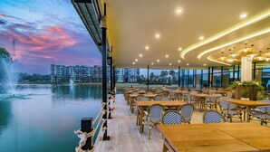 Un restaurante frente al agua con terraza al aire libre, con mesas y sillas de madera y vistas a rascacielos al fondo.