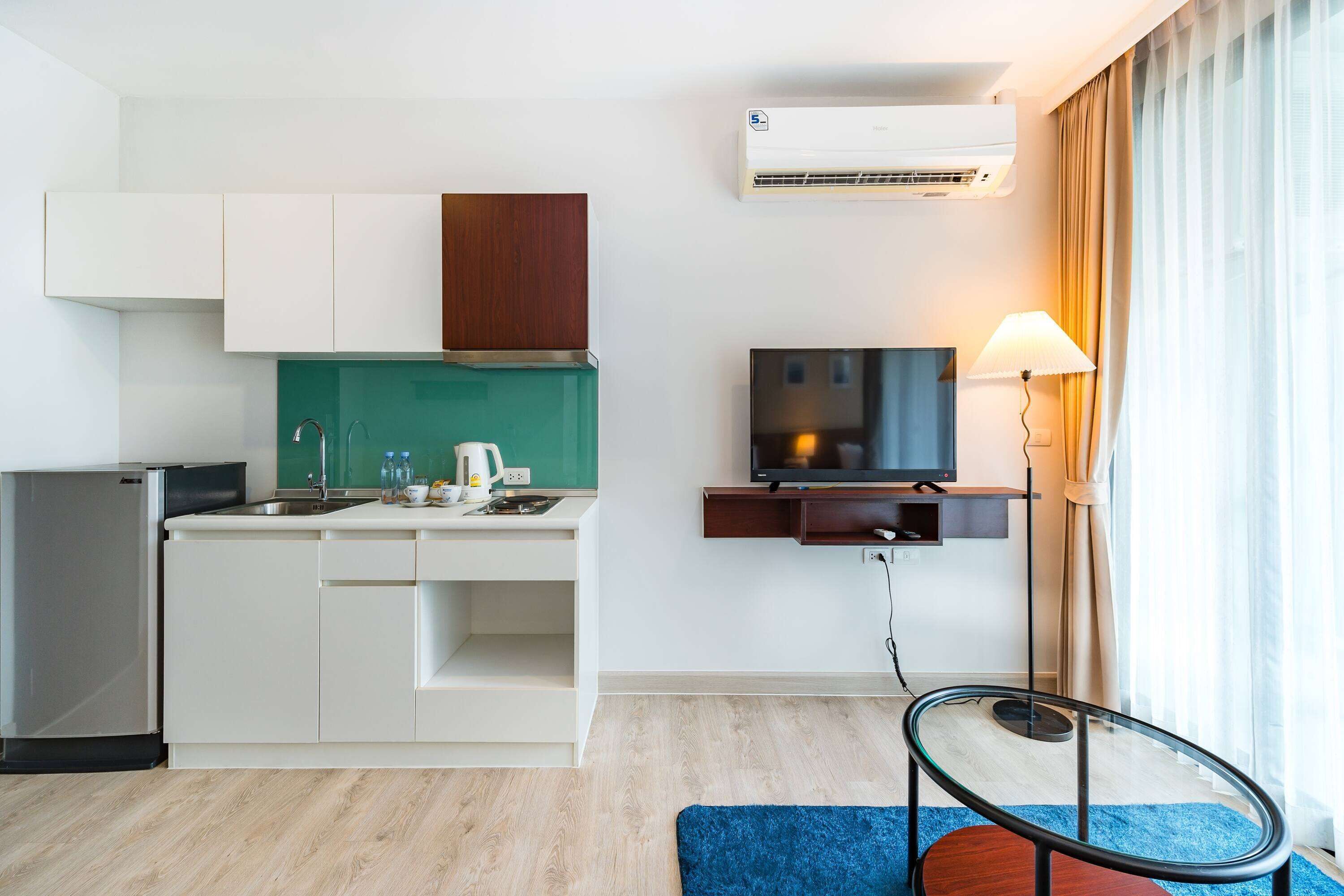 A compact hotel room with a kitchenette, a flat-screen TV, a lamp, and a glass table.