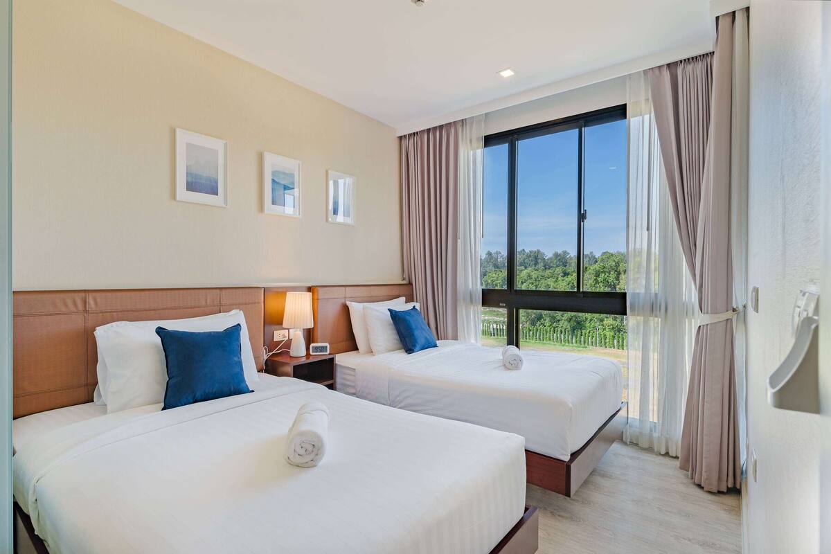 Ein Hotelzimmer mit zwei Betten, einem großen Fenster mit Vorhängen und Blick auf Grünflächen.