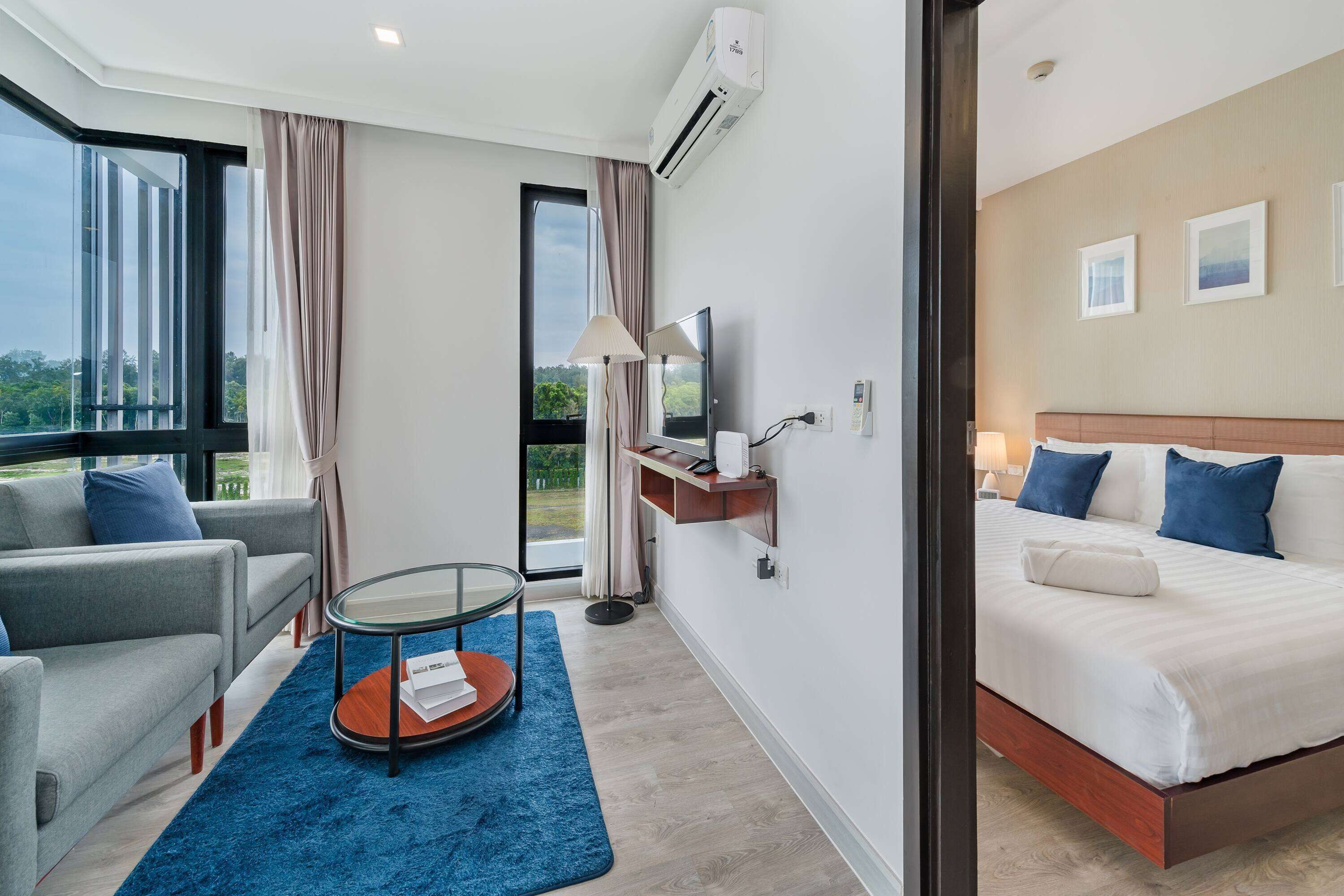 A modern hotel room with a large bed, a sofa, a glass coffee table, and a view of greenery outside.