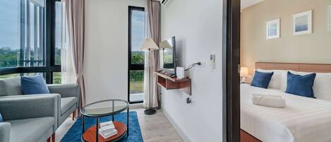 A modern hotel room with a large bed, a sofa, a glass coffee table, and a view of greenery outside.