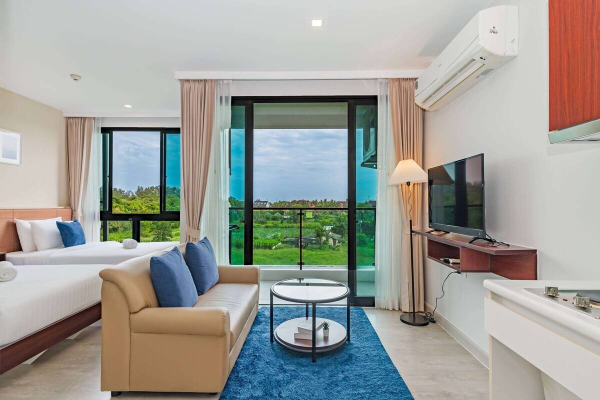 Ein Hotelzimmer mit zwei Betten, einem Sofa, einem Fernseher und einem großen Fenster mit Blick auf Grünflächen.