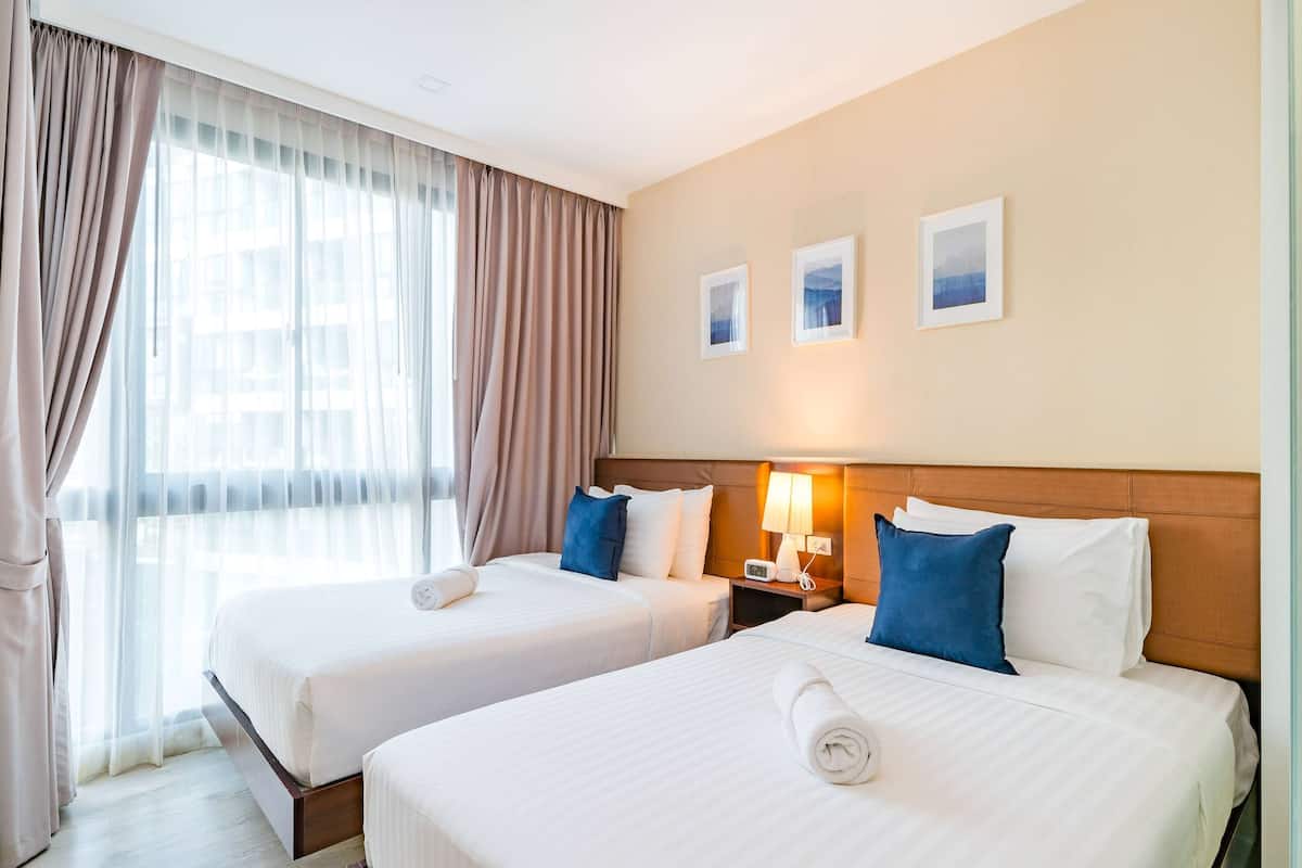 Ein Hotelzimmer mit zwei Betten, einem Nachttisch mit Lampe und einem Fenster mit leichtem Vorhang.