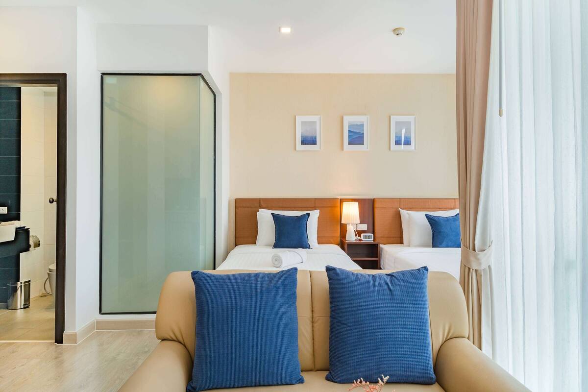 Ein Hotelzimmer mit einem Bett, zwei Nachttischen, einer Ablage, einer blauen Sitzsofa mit Kissen, einer Glastür und einer Wand mit drei gerahmten Bildern.