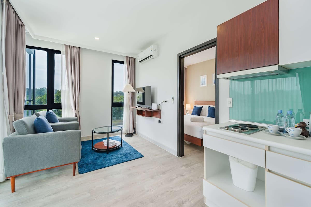 Ein modernes Hotelzimmer mit grauem Sofa, einem kleinen runden Tisch, einem Fernseher, einer Küchenzeile und einem großen Fenster mit Vorhängen.