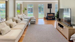 A spacious living room with a large sofa, a fireplace, a TV, and large windows offering a view of the outdoors. - Tallowood House - A great family getaway (NULKABA)