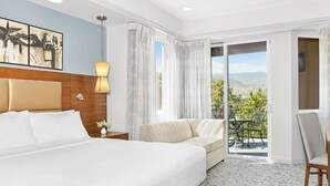 Habitación de hotel con una cama grande, un sofá, un balcón con mesa y sillas, y vistas a las montañas.