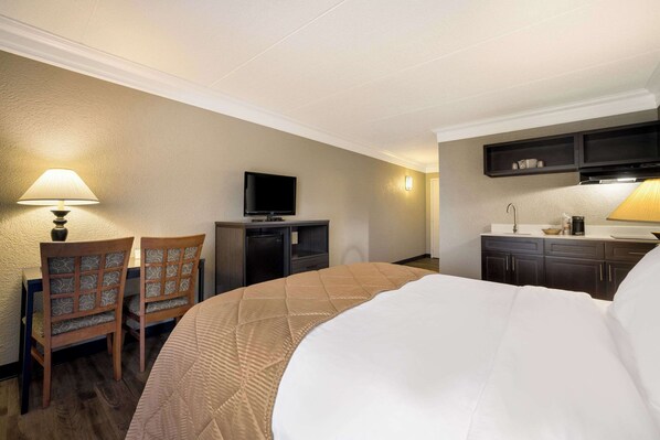 A hotel room with a large bed, a TV, a desk with a lamp, and a kitchenette. - Suburban Studios Syracuse Fairgrounds (Syracuse)