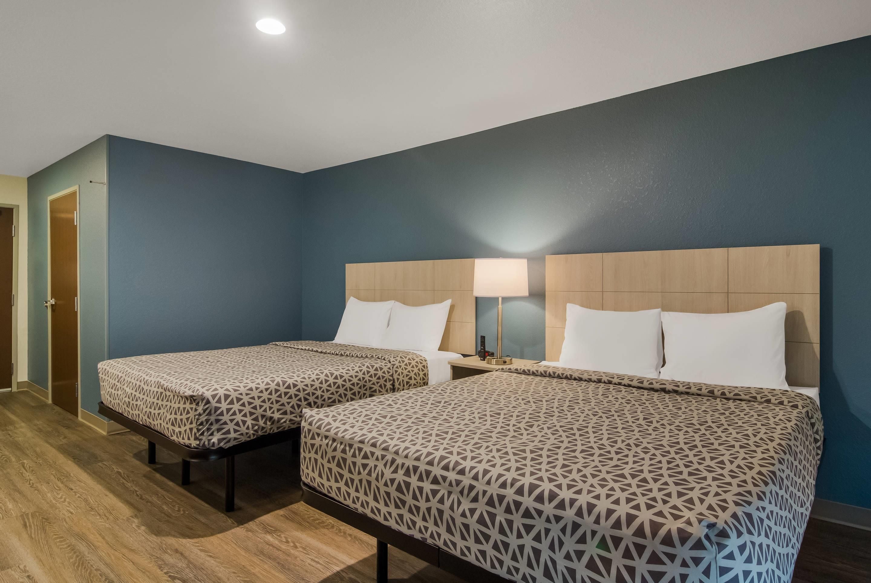Habitación de hotel con dos camas, una mesita de noche, una lámpara y suelo de madera.