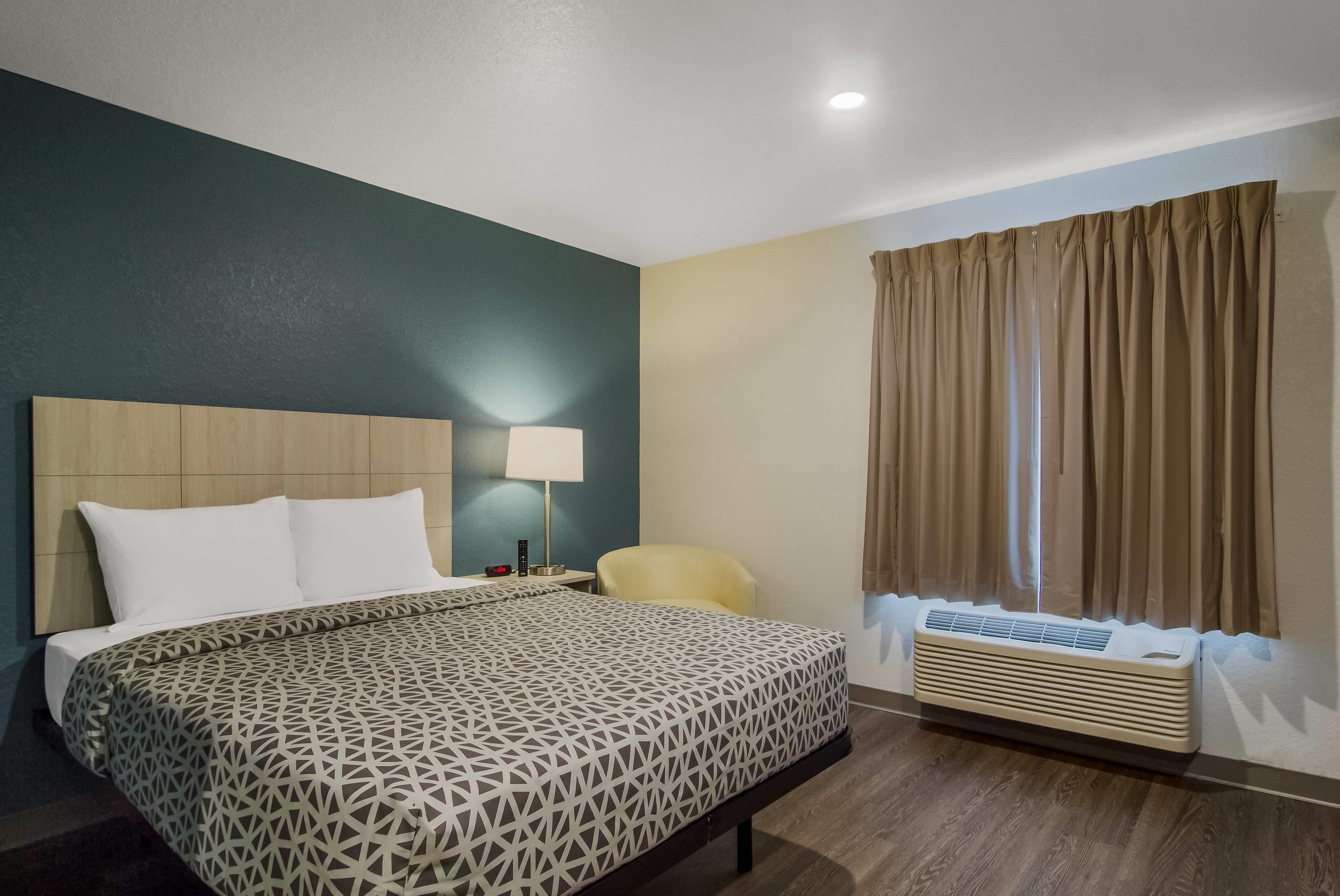 Habitación de hotel con cama, una silla, una lámpara, una mesita de noche y una ventana con cortinas.