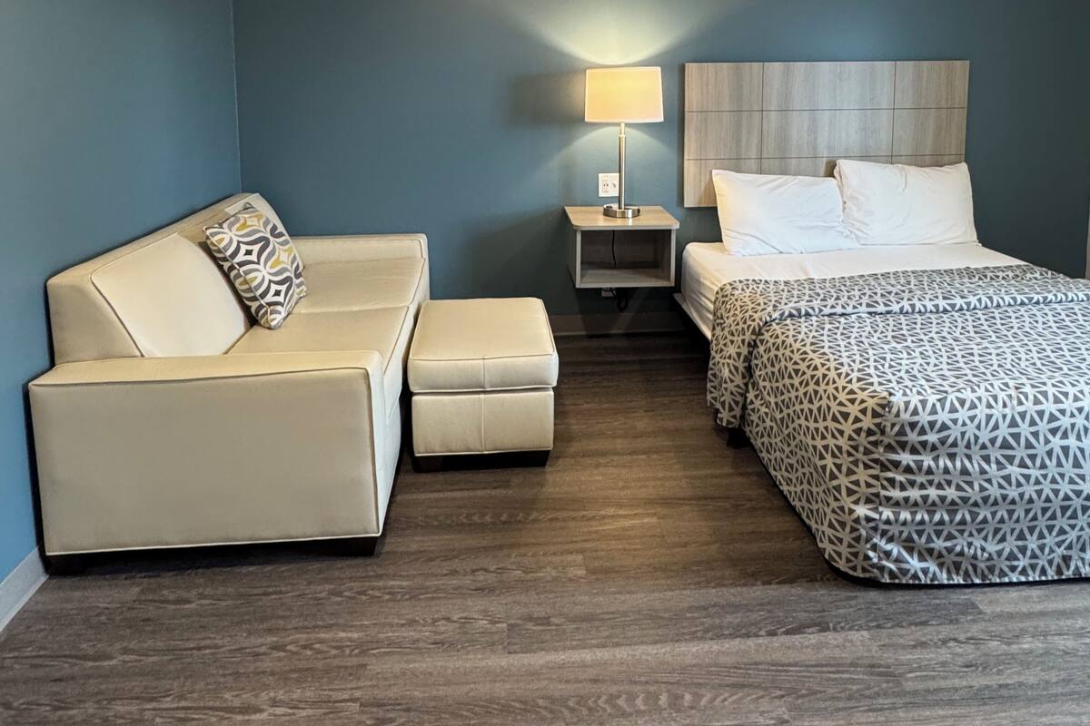 A hotel room with a bed, a sofa, an ottoman, a nightstand with a lamp, and a ceiling light.