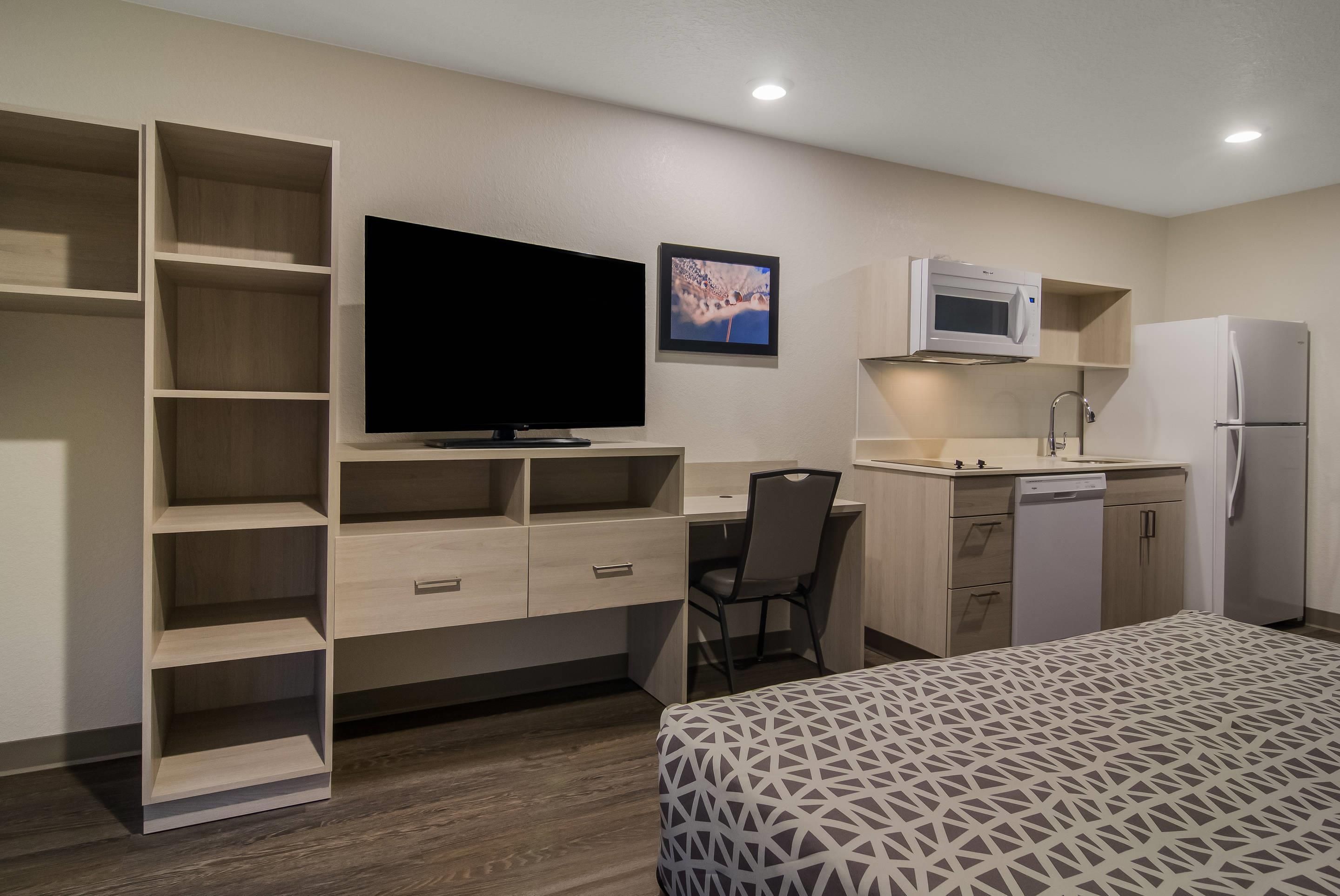 Quarto de hotel com cama, escrivaninha com cadeira, televisão, micro-ondas, frigorífico e estantes embutidas.