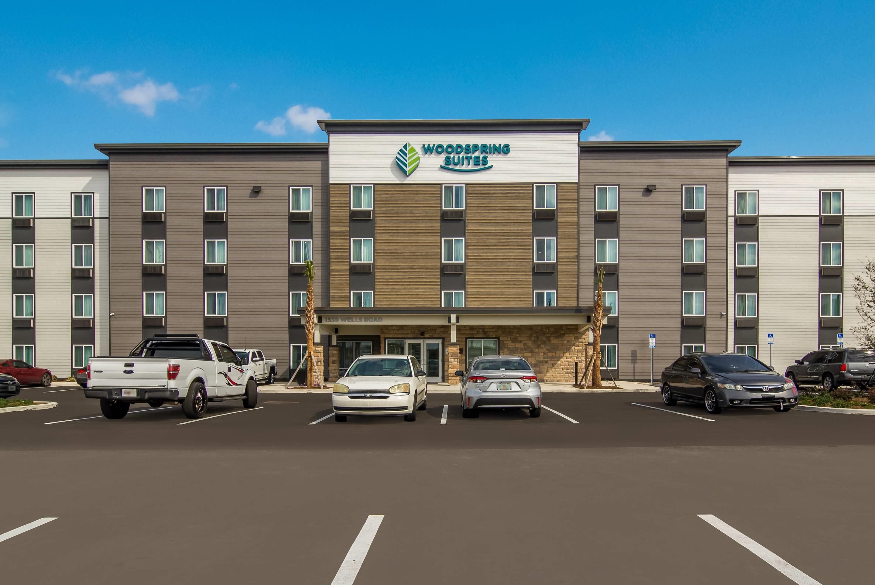 A multi-story hotel with a sign reading 'Woodspring Suites' and a parking lot with several cars.