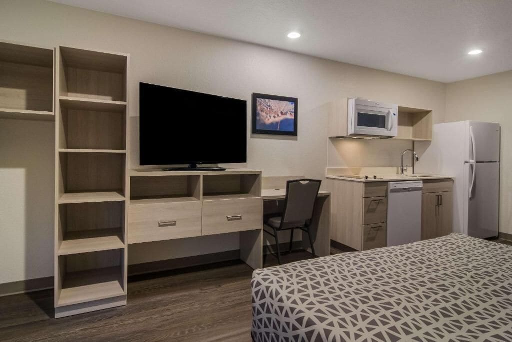 A compact hotel room with a bed, a desk, a chair, a television, a microwave, a refrigerator, and a small kitchenette.