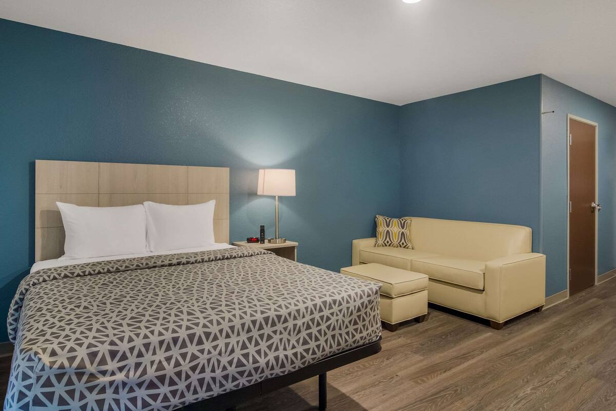 A hotel room with a bed, a bedside table with a lamp, a sofa, and a door.