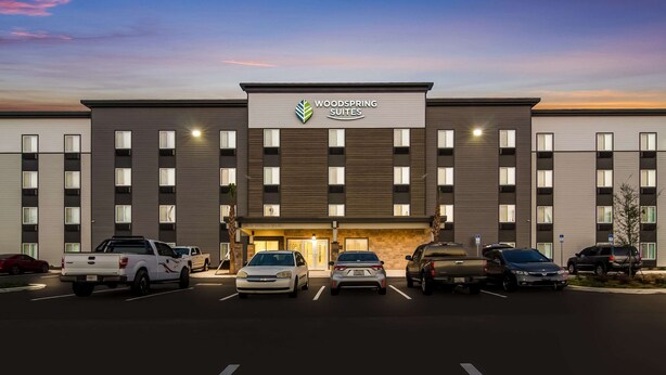 A modern hotel with a flat roof, a sign reading "Woodspring Suites," and a parking lot with several cars.