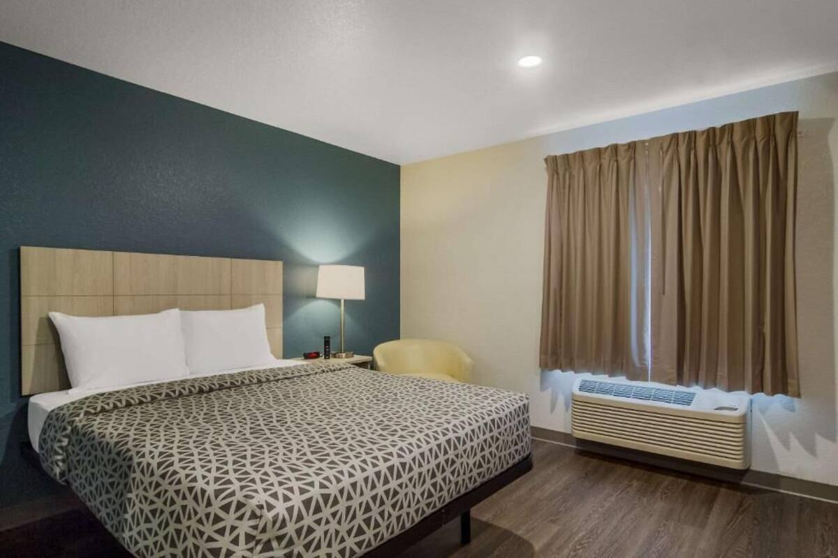 A hotel room with a bed, a chair, a lamp, and an air conditioning unit.