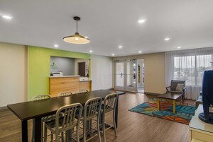 Quarto de hotel moderno com área de jantar, balcão de recepção e uma grande janela com cortinas.