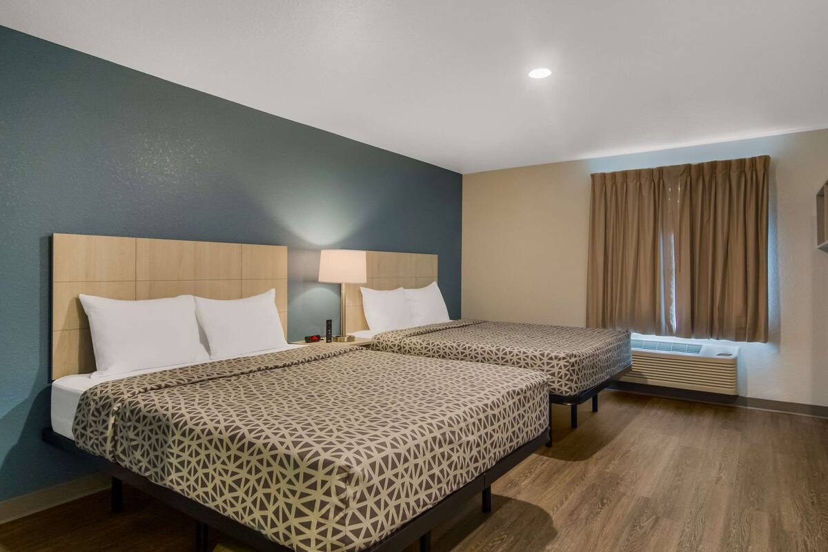 A hotel room with two beds, a nightstand with a lamp, a window with curtains, and an air conditioning unit.