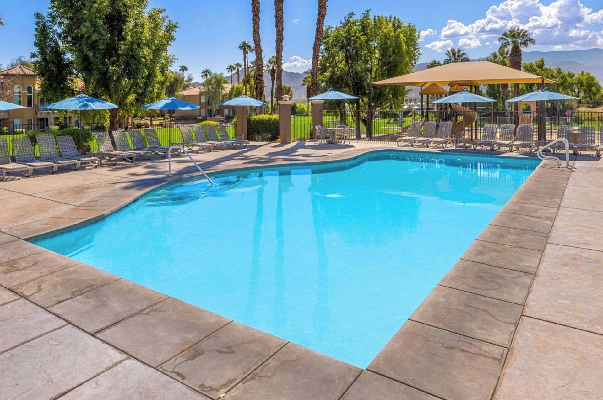Une piscine rectangulaire, bordée d’une terrasse carrelée, entourée de chaises longues et de parasols, avec des palmiers et une vue sur les montagnes en arrière-plan.
