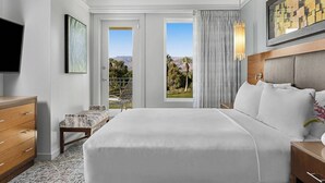 A hotel room with a large bed, a desk, a chair, and a TV. There are two windows with curtains, and a painting on the wall.