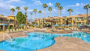 A resort pool with lounge chairs and umbrellas, surrounded by palm trees and hotel buildings.