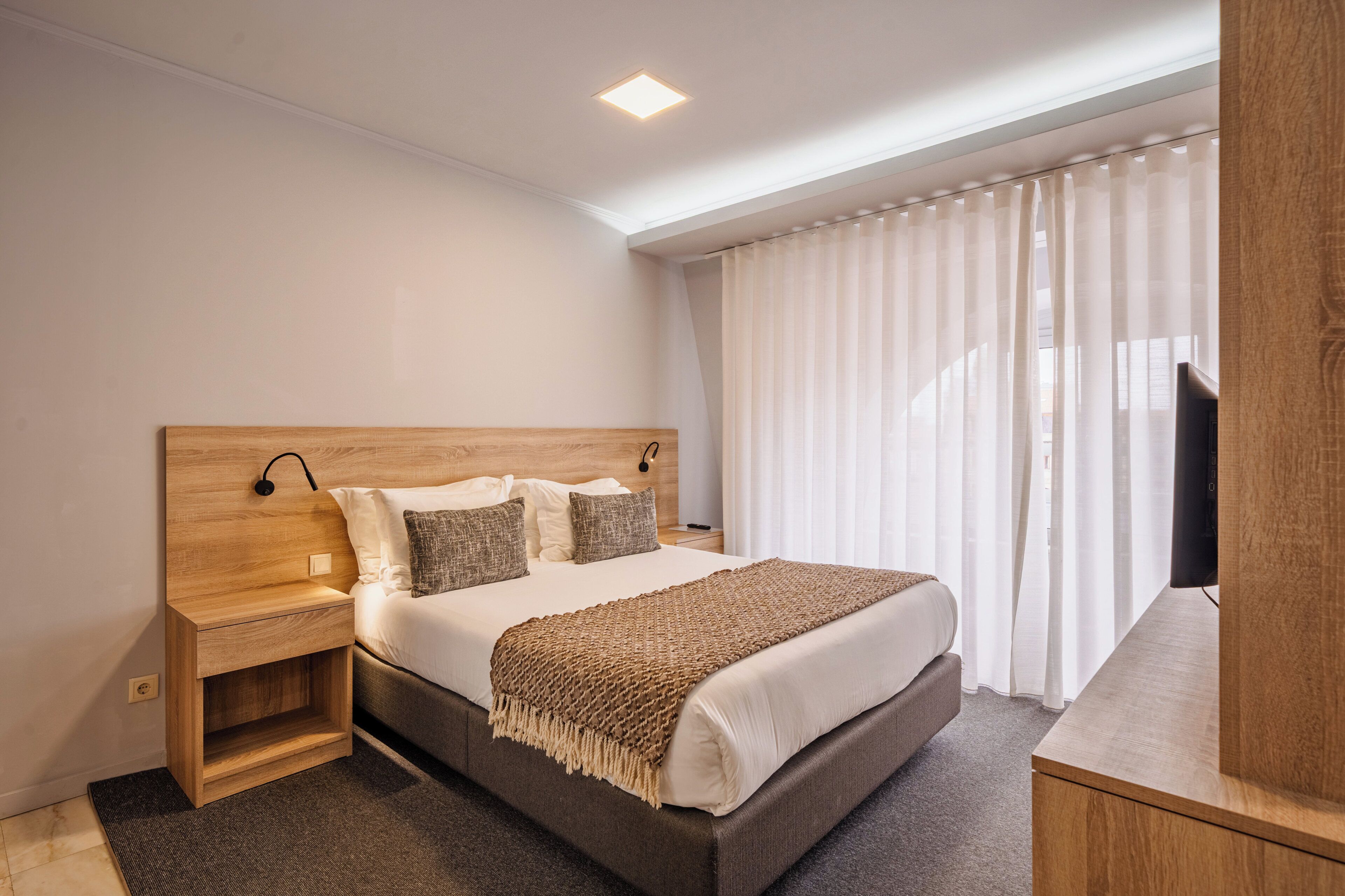 A modern hotel room with a wooden headboard, a large bed with white linens, a bedside table with a lamp, and a TV on a wooden stand.