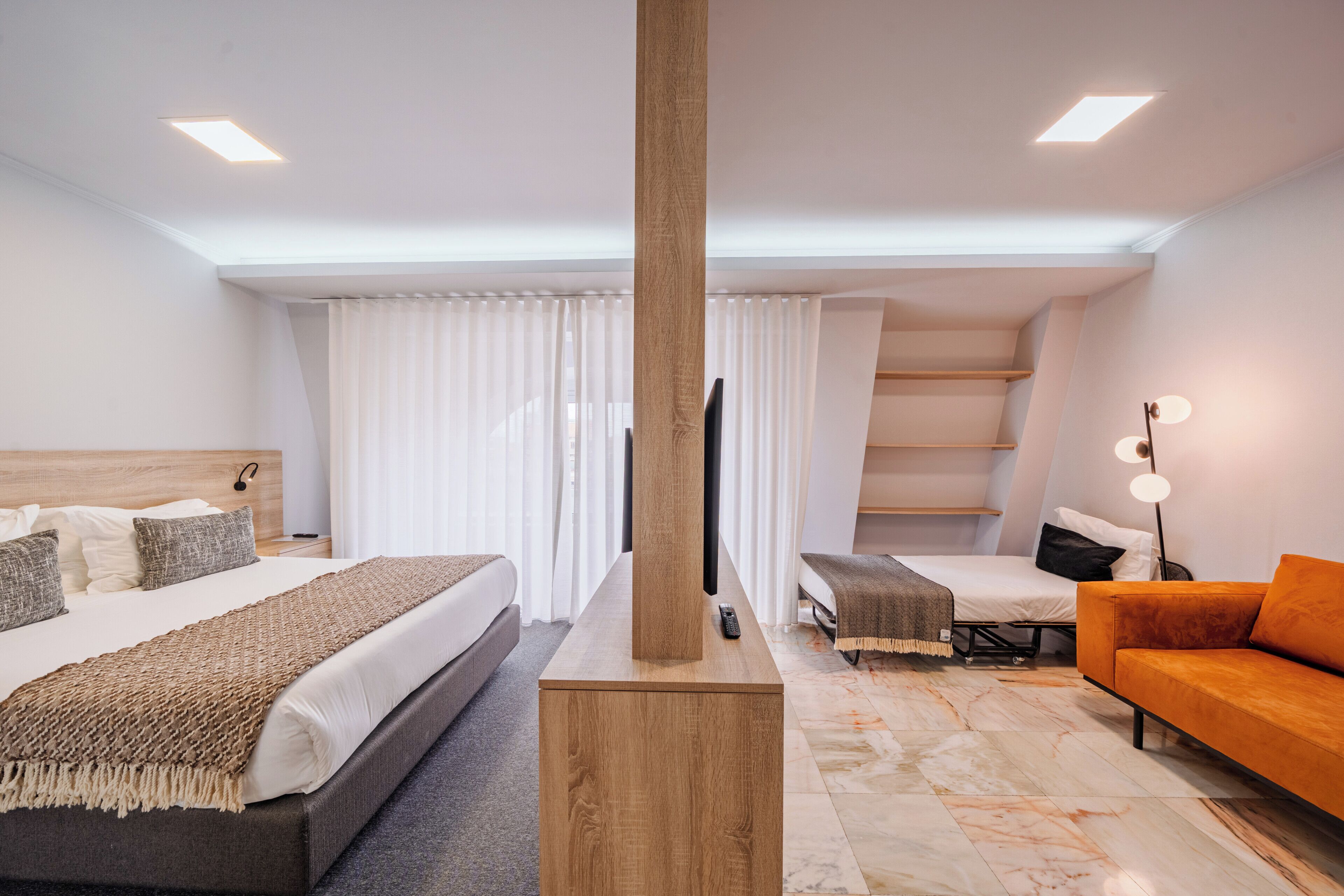 A modern hotel room with a large bed, a sofa, a TV, and a wooden column.