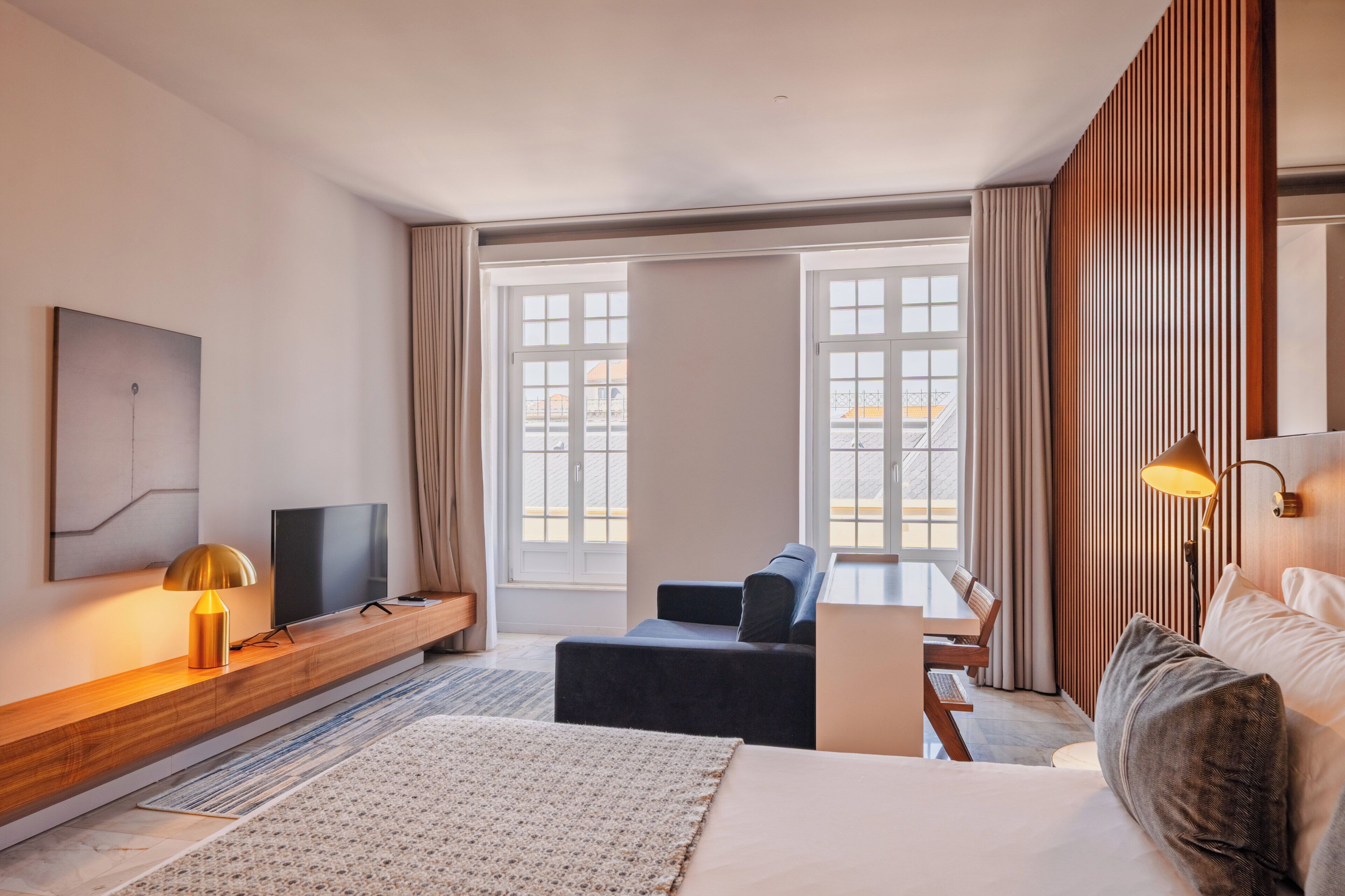 A hotel room with a bed, a desk, a TV, a lamp, and large windows.