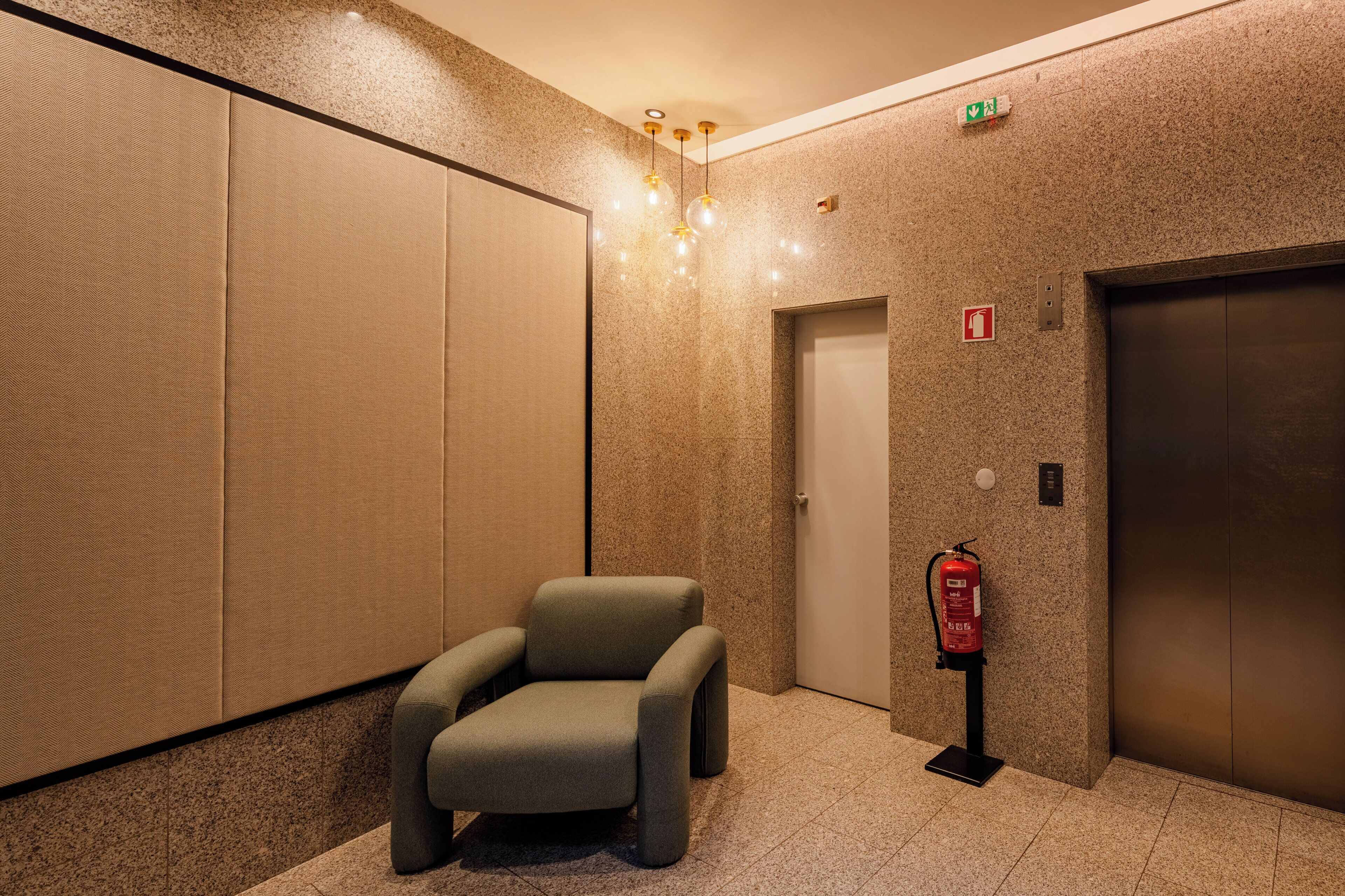 A modern elevator area with a fire extinguisher, an emergency exit sign, and a chair.