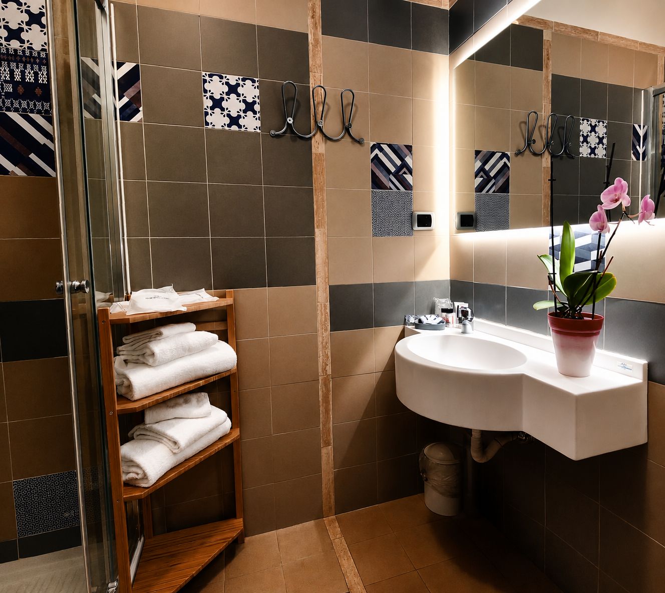 A bathroom with a glass shower enclosure, a wooden towel rack, a white sink with a potted plant, and patterned tiles.