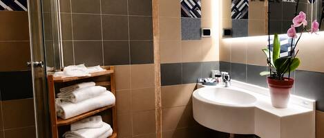 Un baño con cabina de ducha de vidrio, toallero de madera, lavamanos blanco con planta en maceta y azulejos estampados.