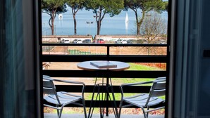 A balcony with two metal chairs and a small round table, overlooking a parking area and a body of water with trees and a sailboat. - HA HOTEL (Bracciano)