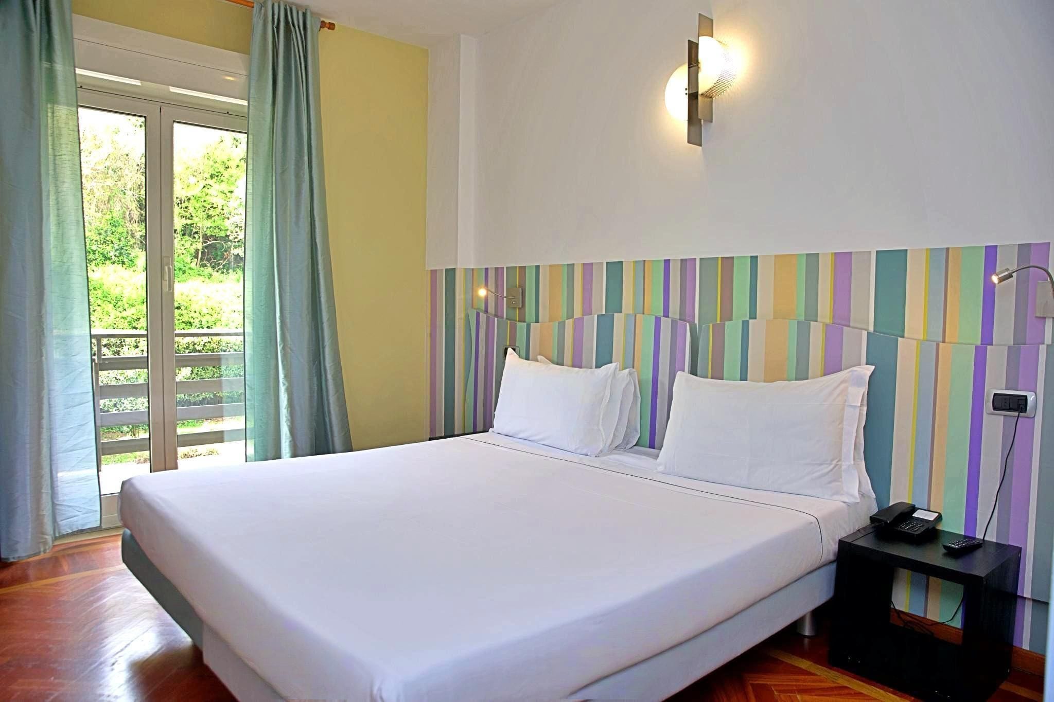 A hotel room with a large bed, a nightstand with a phone, and a view of greenery through the window.