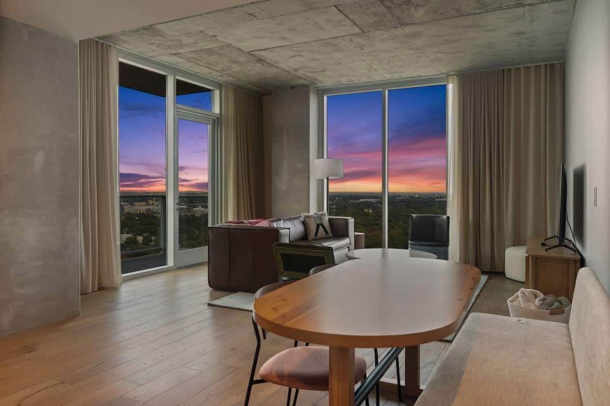 A modern hotel room with a large sliding glass door, a round dining table, a sofa, a TV, and a view of the cityscape at sunset.