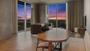 Ein modernes Hotelzimmer mit einer großen Glasschiebetür, einem runden Esstisch, einer Couch, einem Fernseher und Blick auf die Skyline im Sonnenuntergang.