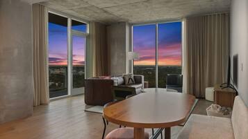 A modern hotel room with a large sliding glass door, a round dining table, a sofa, a TV, and a view of the cityscape at sunset.