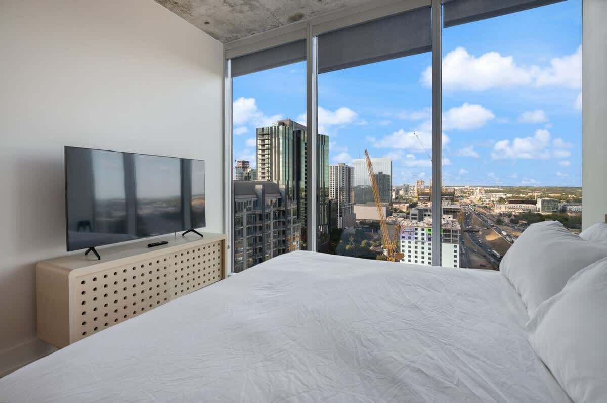 A modern hotel room with a large bed, a television on a wooden console, and a city view through floor-to-ceiling windows.