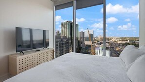 Ein modernes Hotelzimmer mit einem großen Bett, einem Fernseher auf einer Holzkonsole und Blick auf die Stadt durch raumhohe Fenster.