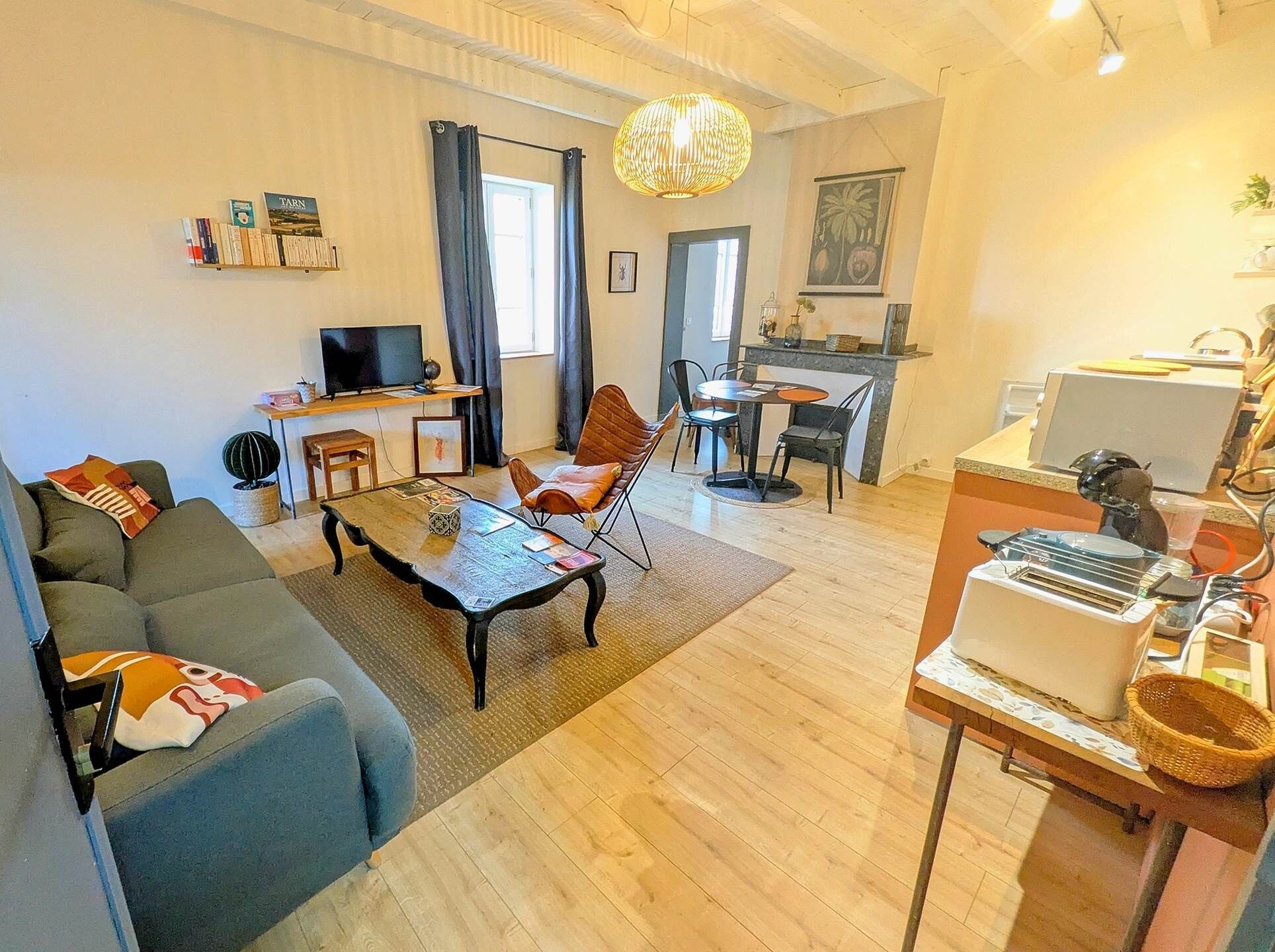 A living room with a sofa, coffee table, and TV. There is a dining area with chairs and a table. A kitchen area with a stove and counter is visible.