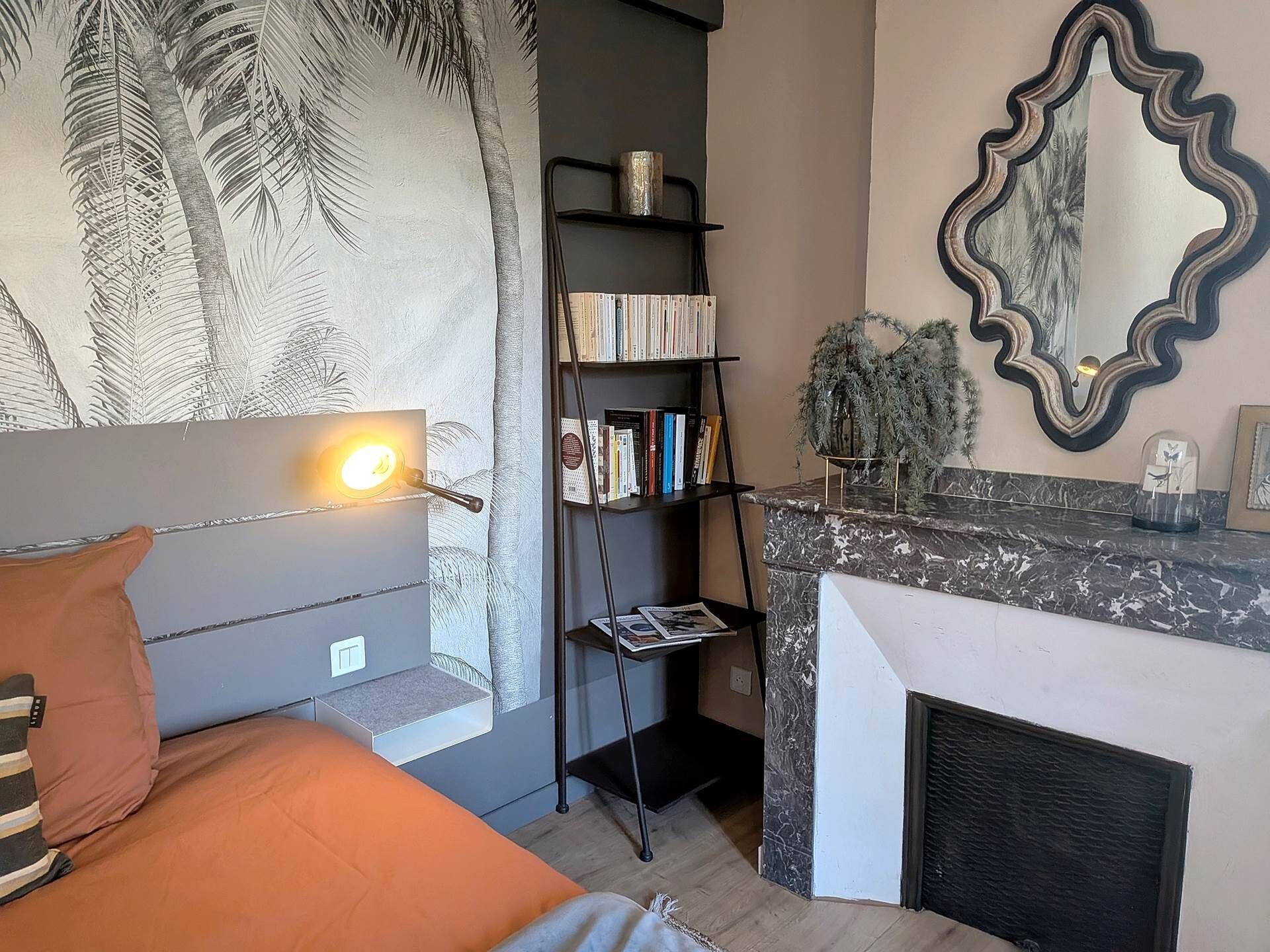 A bedroom with a bed, a bedside lamp, a bookshelf, a mirror, and a fireplace.
